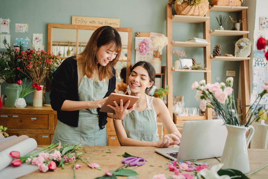 Two women working inside a cozy flower shop with pastel green walls and wooden shelving, surrounded by various fresh flowers including pink roses, white carnations, and red blossoms. One woman with sh