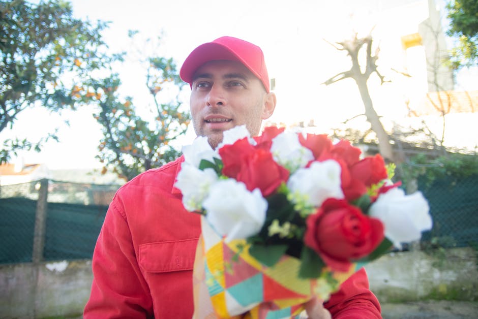 A delivery person dressed in a red uniform and matching cap holds a floral bouquet featuring red and white roses, with green foliage, wrapped in colorful paper. The flowers appear fresh with vibrant p