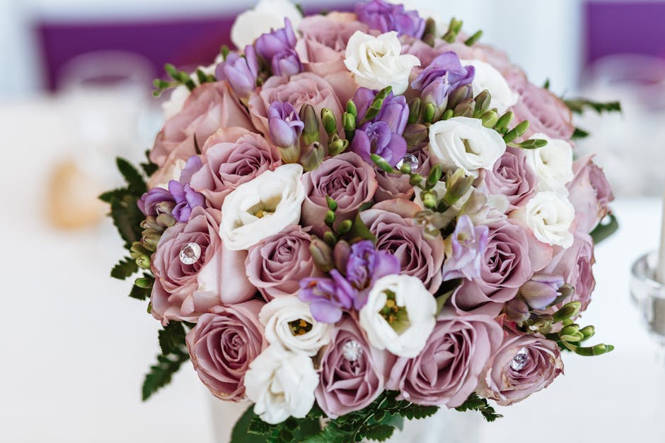 A close-up of a floral arrangement featuring a bouquet of fresh lavender, pale pink roses, and white lisianthus flowers, accented with small purple freesia buds and green foliage. The bouquet is arran