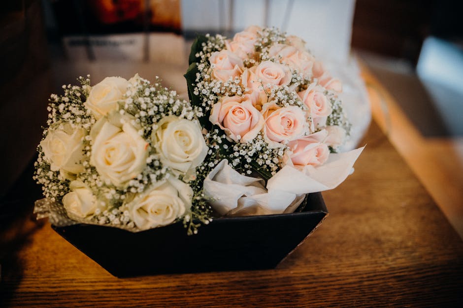 A black rectangular box on a wooden surface contains two bouquets of fresh flowers, one with creamy white roses and the other with soft pink roses, both accented with delicate white baby's breath. The